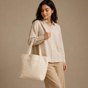 Pode incluir: Uma camisa bege de manga comprida com botões e calças castanho-claras, combinadas com um saco tote grande e retangular de lona em branco sujo, com duas alças resistentes e um bolso exterior com detalhes de costura vertical.