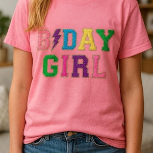May include: Pink t-shirt with the text "B-DAY GIRL" in colorful, glittery letters. The shirt is a crew neck style and is paired with blue jeans. The overall look is casual and festive.
