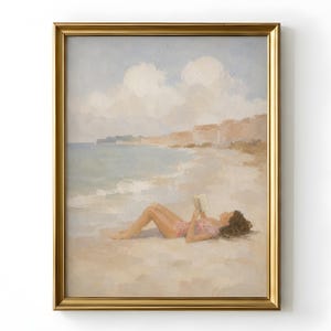 May include: A framed oil painting depicting a person lying on a sandy beach reading a book. The scene features a calm ocean, soft clouds in a pale blue sky, and distant cliffs. The artwork is enclosed in a metallic gold rectangular frame.
