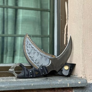 May include: A curved karambit knife with a black and wood-grain handle and a silver blade. The knife rests on a black leather sheath with a gold button. The blade has a textured pattern and is partially covered by a decorative leather piece.