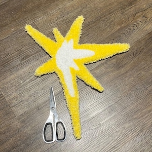 May include: A handmade, star-shaped rug with a bright yellow exterior and a white interior design. The rug is placed on a wooden floor next to a pair of silver scissors.