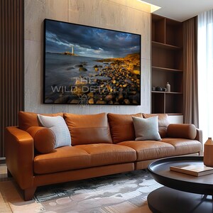May include: A large framed canvas print depicting a coastal scene with a lighthouse under a dramatic, cloudy sky. The artwork is displayed above a brown leather sofa with decorative pillows. The text "THE WILD DETAIL" is visible on the print.