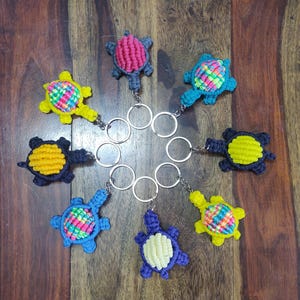 Puede incluir: Una colección de llaveros de macramé hechos a mano con forma de tortuga en varios colores, incluyendo amarillo, azul y rosa. Cada llavero presenta un diseño de tortuga con un llavero de metal adjunto. Las tortugas están dispuestas sobre una superficie de madera.