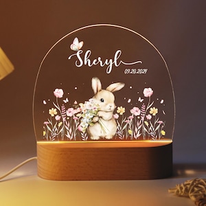 May include: An arched acrylic night light with a wooden base featuring a printed illustration of a rabbit holding a bouquet of flowers, a butterfly, and the text Sheryl and 09.20.2021. The design includes pink and yellow wildflowers on stems.
