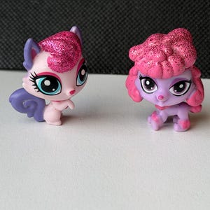 May include: Two small, colorful toy figurines. One is a pink and purple cat-like creature with glittery pink hair and blue wings. The other is a purple poodle-like figure with pink glittery hair. Both have large, expressive eyes.