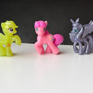 May include: Three colorful toy ponies are displayed on a white surface. The ponies are in shades of yellow, pink, and purple. The yellow pony has wings, the pink pony has a crescent moon design, and the purple pony has wings and a horn.
