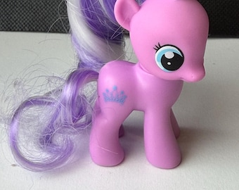 May include: A pink toy pony with a purple and white mane and tail. The pony has blue eyes and a crown design on its side. The toy is made of plastic and has a glossy finish. The pony is standing on a white surface.