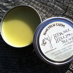 May include: A small tin container of Honeyrun Farm Herbal Healing Salve with Propolis. The label is white with black text and a small illustration of a plant. The text on the label reads "Honeyrun Farm", "Herbal Healing Salve", "With Propolis", "Apply to cuts, scrapes, bruises, insect bites, and dry skin", "Ingredients: Olive Oil (infused with Propolis, Mullein, and Comfrey), Beeswax, Tea Tree Essential Oil".