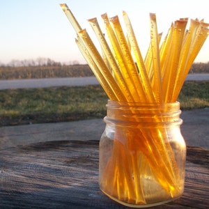May include: A glass jar filled with honey sticks. The sticks are a light amber color and are arranged vertically in the jar. The jar is sitting on a wooden surface.