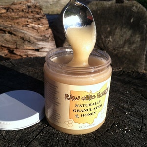 May include: A glass jar of raw Ohio honey with a white label that reads "Raw Ohio Honey Naturally Granulated Honey 13 oz (369g)". A silver spoon is dipping into the honey, which is a light golden color.