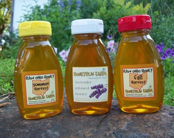 May include: Three glass bottles of raw Ohio honey with yellow, white, and red lids. The labels are for summer harvest, lavender infused honey, and fall harvest. The bottles are sitting on a rock.