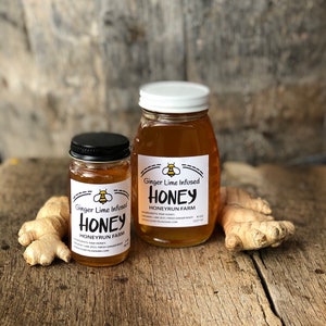 May include: Two glass jars of honey with white and black labels. The labels say "Ginger Lime Infused Honey" and "Honeyrun Farm". The jars are sitting on a wooden surface with fresh ginger root.