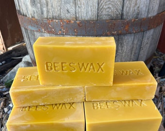 May include: A stack of five yellow beeswax blocks with the word "BEESWAX" embossed on each block. The blocks are stacked on a wooden surface in front of a wooden barrel.