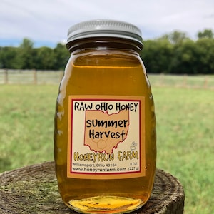 May include: A glass jar of raw Ohio honey labeled "Summer Harvest" from Honeyrun Farm in Williamsport, Ohio. The jar is 8 ounces (227 grams) and is sitting on a tree stump.