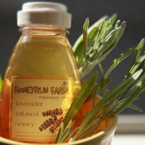May include: A clear glass bottle of honey infused with lavender. The bottle has a white label with the text "Honeyrun Farm, Williamsport, Ohio, lavender infused honey, www.honeyrunfarm.com".