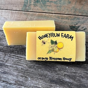 May include: Two bars of yellow soap with a label that reads "Honeyrun Farm Orange Blossom Honey". The label features a cartoon bee and orange blossoms.
