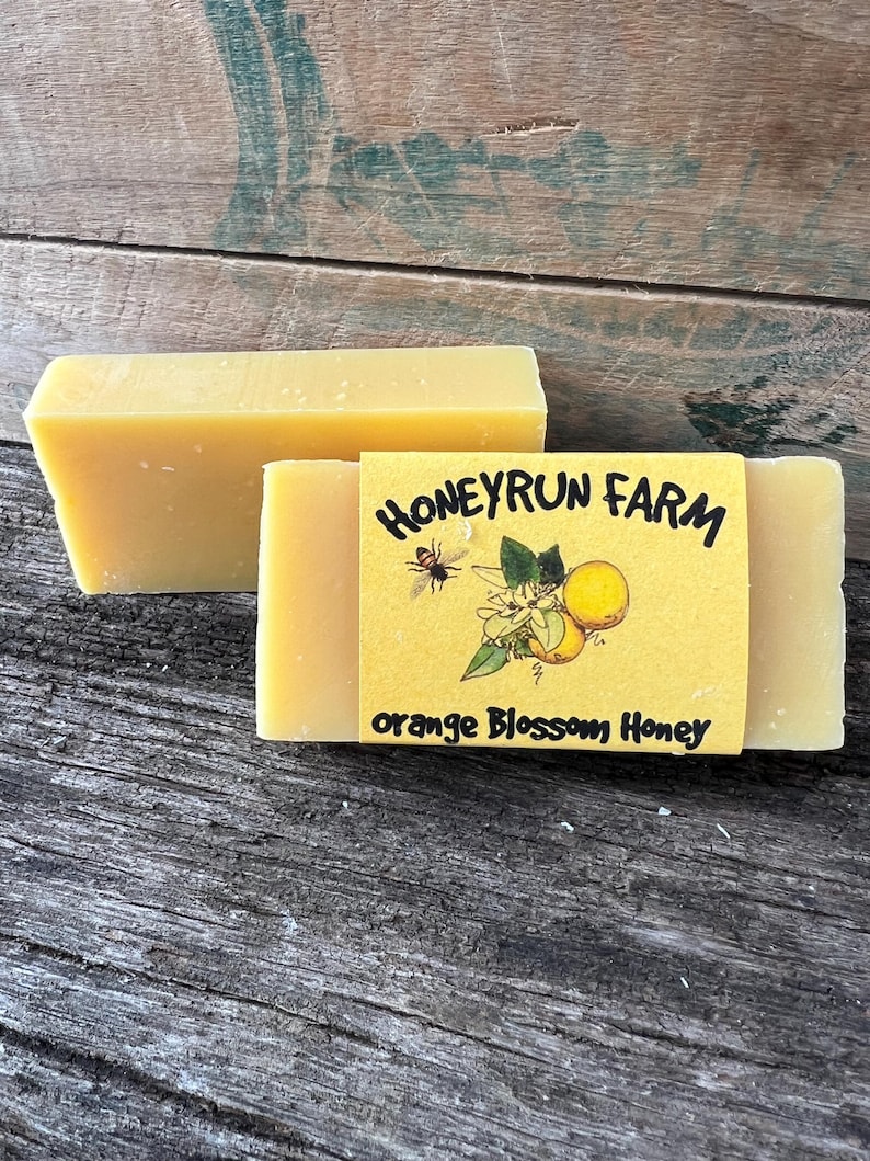 May include: Two bars of yellow soap with a label that reads "Honeyrun Farm Orange Blossom Honey". The label features a cartoon bee and orange blossoms.