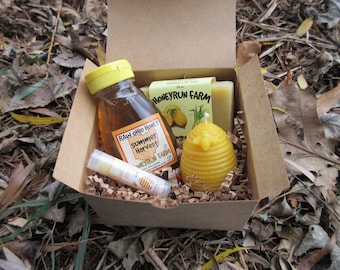 May include: A brown cardboard box filled with a honey gift set. The set includes a small bottle of raw honey, a lip balm, a bar of soap, and a beeswax candle shaped like a honeycomb.