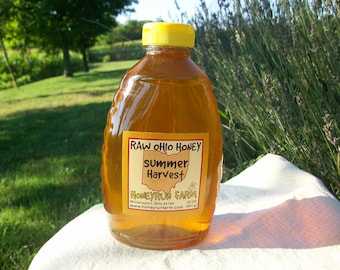 Puede incluir: Una botella de vidrio transparente de miel cruda de Ohio con una tapa amarilla. La etiqueta dice "RAW OHIO HONEY", "Summer Harvest", "HONEYRUN FARM", "Williamsport, Ohio 43164", "www.honeyrunfarm.com" y "32 oz".