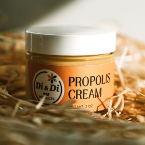 May include: A jar of propolis cream with a white lid and an orange label. The label features the text "PROPOLIS CREAM" and the brand name "Di & Di BEE PRODUCTS". The jar is set on a bed of straw and the product weighs 2 oz.