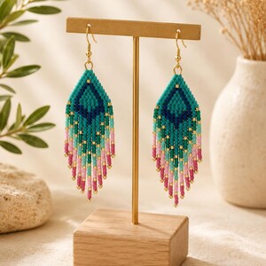 Peut inclure: Une paire de boucles d'oreilles pendantes avec un motif en forme de losange composé de petites perles en verre dans des tons bleu sarcelle, bleu foncé, vert clair et rose. Elles présentent une longue frange avec des crochets dorés.