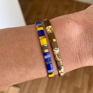 Peut inclure: Deux bracelets de perles. L'un présente un motif de perles rectangulaires jaunes, bleues et bleu clair. L'autre bracelet a un motif de perles rectangulaires marron et blanches avec des accents métalliques dorés. Bijoux.