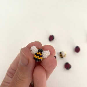 Peut inclure: Une petite épingle à abeille perlée avec des ailes blanches, un corps jaune et noir et un dos d'épingle doré. Plusieurs autres petites épingles à insectes perlées sont éparpillées en arrière-plan, dont des coccinelles. L'épingle à abeille est tenue dans la main d'une personne.