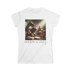 May include: White t-shirt featuring a detailed graphic of a dinosaur reading a book amidst stacks of books. The image includes the text "Unearth a story" in a handwritten style font. The design is centered on the shirt.