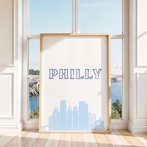 May include: A framed art print featuring the word "PHILLY" in blue lettering above a light blue silhouette of the Philadelphia skyline. The artwork is displayed in a room with a window overlooking a coastal view.