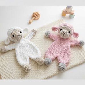 Könnte beinhalten: Zwei gehäkelte Babyspielzeuge in Lammform. Eines ist weiß, das andere rosa. Beide haben beige Gesichter, Ohren und Füße mit grauen Akzenten an Füßen und Händen. Sie liegen auf einer cremefarbenen Decke, mit einer Holzrassel und Spielzeugblöcken im Hintergrund.