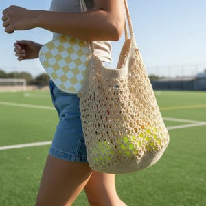 May include: A beige crocheted net tote bag filled with yellow tennis balls. A small, white and yellow checkered purse is also visible. The bags are carried over the shoulder. The background is a green field.