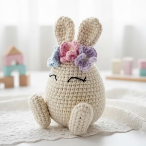 May include: A handmade crocheted bunny doll. The off-white bunny has long ears and a floral crown with pink, purple, and blue flowers. The bunny's face has closed eyes and a smiling mouth. The doll is sitting on a white lace surface.