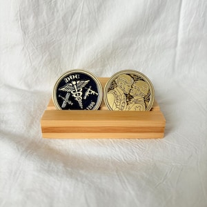 May include: Two gold-colored commemorative coins displayed on a bamboo stand. One coin features the text "DOC" with medical symbols and the year "1890". The other coin depicts two figures in military attire.