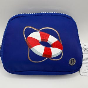 May include: A navy blue zippered pouch with a hand-painted red and white life preserver design. The pouch has a silver zipper pull and a small silver logo. The pouch is designed to hold small items.