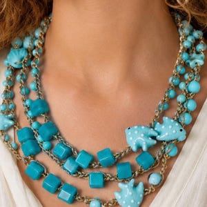 May include: A multi-strand necklace featuring turquoise blue beads in various shapes, including smooth cubes, round spheres, and three distinct fish-shaped charms with white polka dots, all connected by metallic gold-toned chain links.