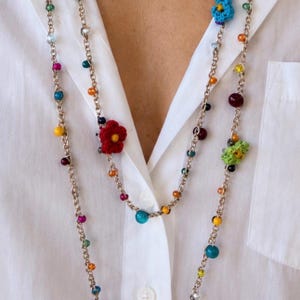 Può includere: Una lunga collana a doppio filo caratterizzata da una catena metallica decorata con varie perline colorate e piccoli accenti floreali all'uncinetto in rosso, blu e verde. La collana è indossata sopra una camicia bianca con bottoni.