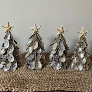 以下が含まれることがあります： 貝殻で作られた6つの装飾的なクリスマスツリー。各ツリーは円錐形で、虹色の貝殻でできており、ヒトデが飾られています。織り込まれたマットの上に配置されています。