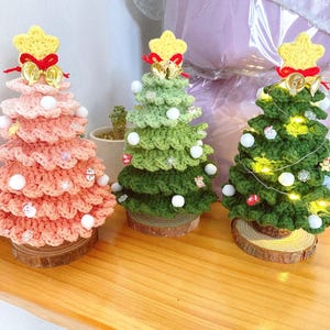 Könnte beinhalten: Drei handgefertigte gehäkelte Weihnachtsbäume in Rosa, Grün und Dunkelgrün, jeweils mit gelbem Sternaufsatz und dekorativen Ornamenten. Die Bäume stehen auf Holzsockeln, der dunkelgrüne Baum mit Lichterketten.