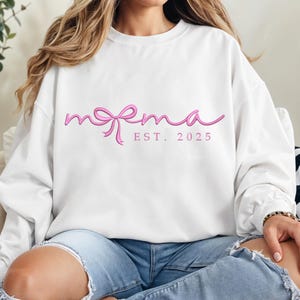 Puede incluir: Sudadera blanca con la palabra "mema" en rosa, con un diseño de lazo. Debajo de la palabra está "EST. 2025" en rosa. La sudadera es de estilo casual, perfecta para el uso diario.