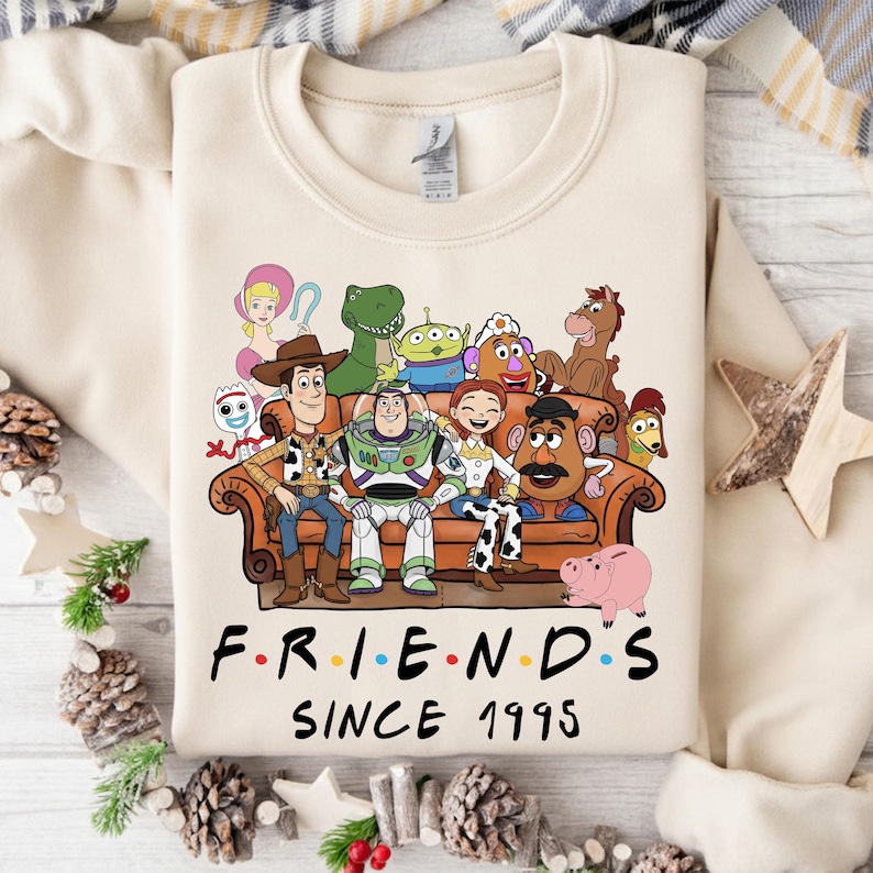 May include: Cream-colored sweatshirt featuring a graphic of characters from the movie Toy Story sitting on a brown couch. The text "F.R.I.E.N.D.S Since 1995" is printed below the graphic in a colorful font. The sweatshirt is a crew neck style.