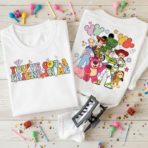 May include: Two white t-shirts with colorful graphics. One shirt has the text "YOU'VE GOT A FRIEND IN ME" in retro font. The other shirt features characters from the Toy Story movie. Party decorations are scattered around the shirts.