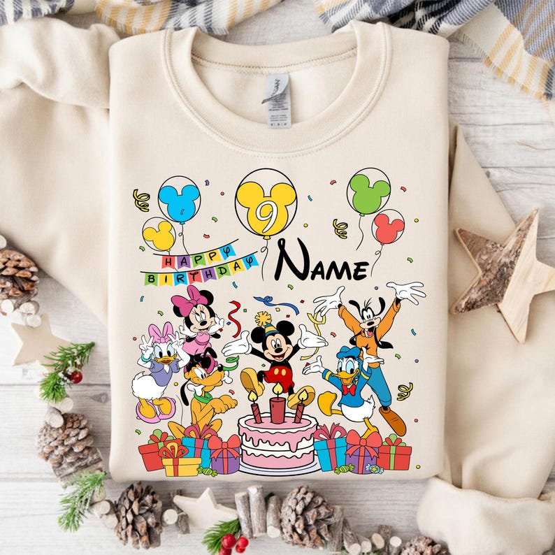 May include: Cream-colored sweatshirt featuring a colorful graphic of Mickey Mouse and friends celebrating a birthday. The design includes balloons, a cake, wrapped gifts, and the text "Happy Birthday" and "Name".