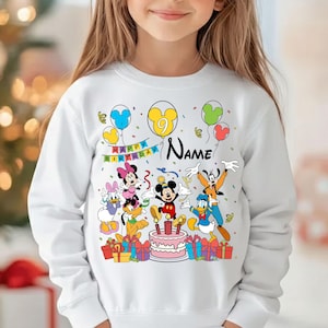 May include: White long-sleeved sweatshirt featuring a colorful Disney-themed birthday design. The design includes Mickey Mouse, Minnie Mouse, Donald Duck, and Goofy, along with balloons, a cake, and the text "Happy Birthday" and "Name".