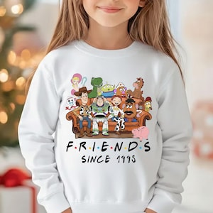 May include: White long-sleeved sweatshirt featuring a colorful graphic of Toy Story characters sitting on a brown couch. The text "FRIENDS SINCE 1995" is printed below the graphic in black.