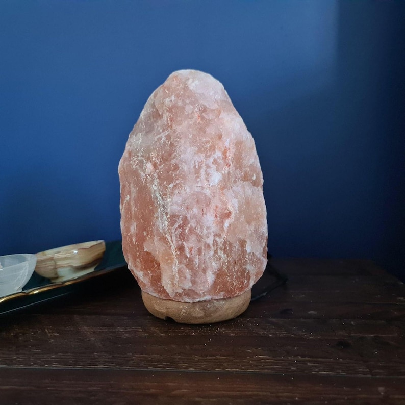 Peut inclure: Une lampe de sel rose de l'Himalaya avec une base en bois. La lampe a une surface rugueuse et textur&eacute;e et est &eacute;clair&eacute;e de l'int&eacute;rieur. L'arri&egrave;re-plan est un mur bleu fonc&eacute;, et la lampe repose sur une surface en bois fonc&eacute;.