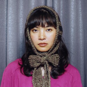 Puede incluir: Un gorro de ganchillo marrón con un detalle de lazo, usado con una camisa rosa. El gorro enmarca la cara y cae por delante, creando un look elegante. La camisa rosa presenta un diseño gráfico en la parte delantera.