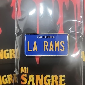 Könnte beinhalten: Ein blau-goldener LA Rams-Kennzeichen-Pin. Der Pin trägt oben "CALIFORNIA" und unten "LA RAMS". Der Pin ist auf einem schwarzen Hintergrund mit den Worten "MI SANGRE" in Gelb verpackt.