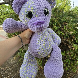 Pode incluir: Um urso de peluche feito em croché com fio grosso e macio de cor lavanda com manchas amarelas e verdes. O urso apresenta grandes olhos de segurança pretos brilhantes, um nariz roxo bordado e orelhas arredondadas, com um padrão de pontos texturizados.