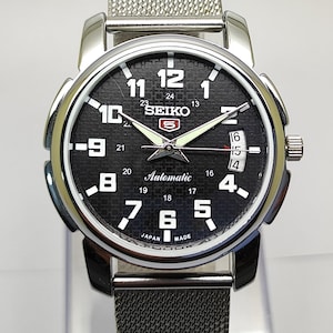 Op de afbeelding: Een zilverkleurig Seiko 5 automatisch horloge met een zwarte wijzerplaat. Het horloge heeft witte cijfers, een datumweergave en een mesh band. De wijzerplaat heeft de tekst "SEIKO" en "Automatic".