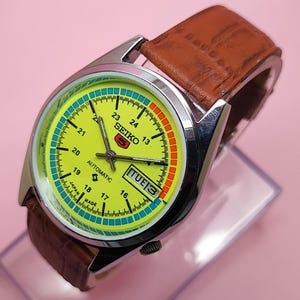 Może przedstawiać: Zegarek automatyczny Seiko 5 w stylu vintage z żółtą tarczą, srebrną kopertą i brązowym skórzanym paskiem. Tarcza posiada czarne cyfry, wyświetlacz dnia i daty oraz logo Seiko. Zegarek jest prezentowany na przezroczystym stojaku.